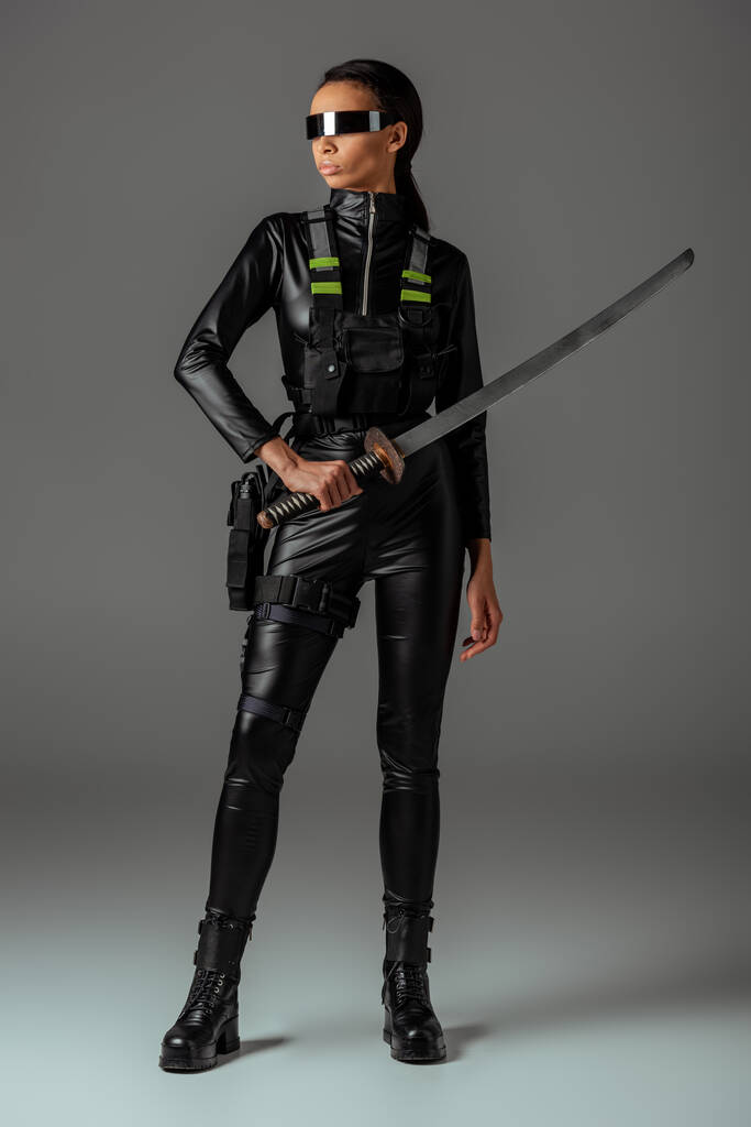 aantrekkelijke futuristische Afrikaans-Amerikaanse vrouw in bril met zwaard op grijs - Foto, afbeelding