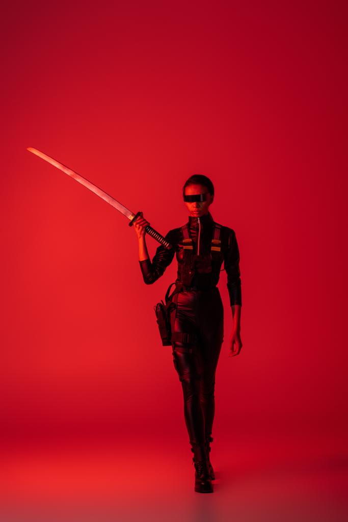 futuristic african american woman in glasses walking with sword on red background - Photo, Image