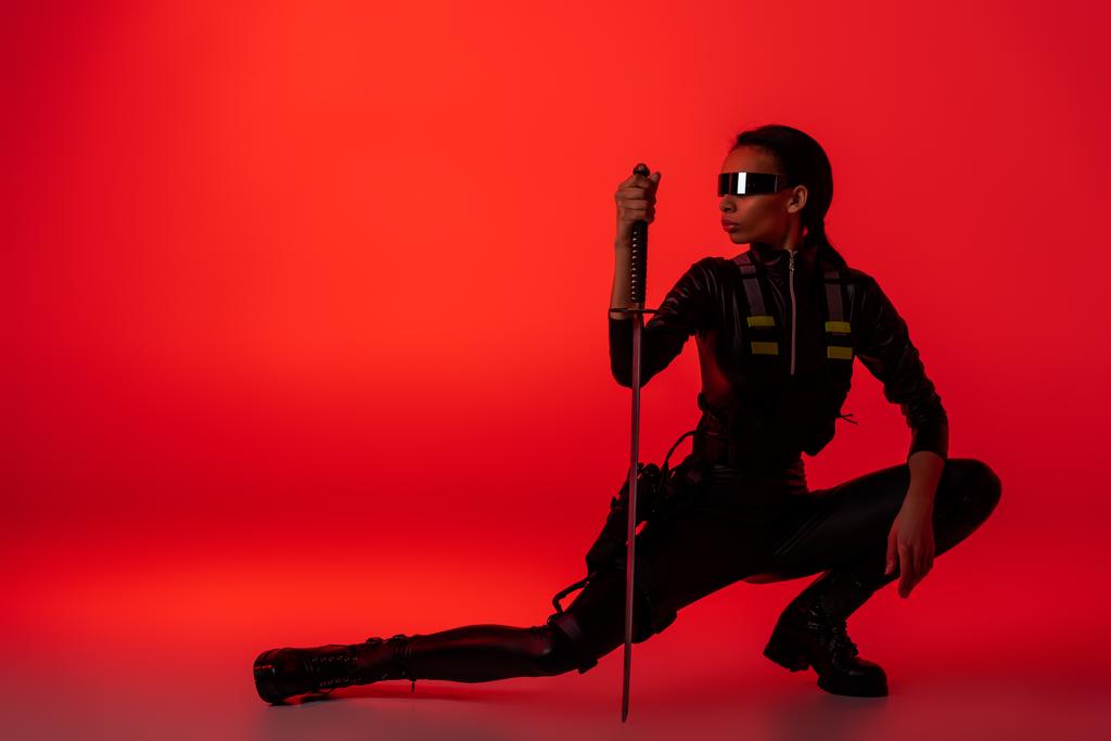 φουτουριστική Αφροαμερικανή γυναίκα με γυαλιά κάθεται με το σπαθί στο κόκκινο φόντο - Φωτογραφία, εικόνα