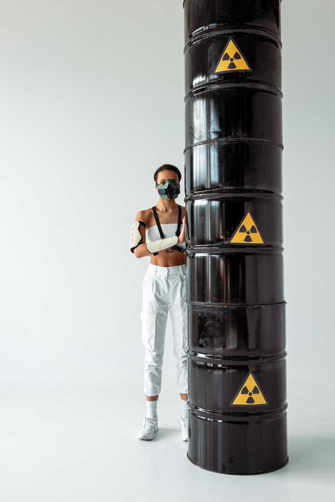futuristic african american woman in safety mask near radioactive waste barrels on white background - Photo, Image