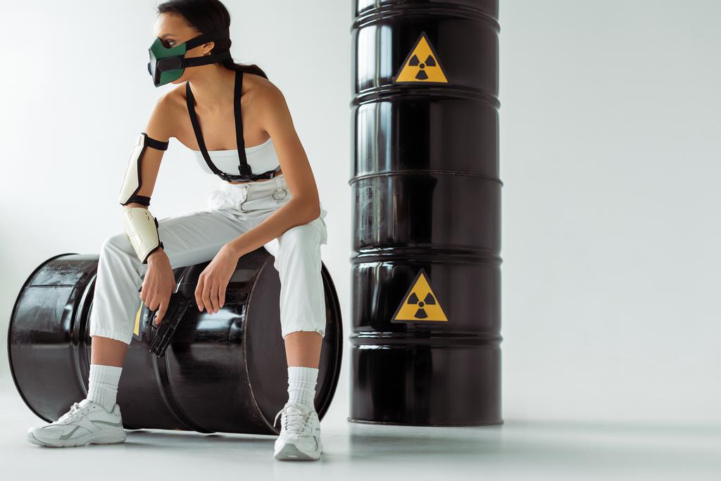 футуристическая американка в защитной маске с пистолетом возле бочек с радиоактивными отходами на белом фоне
 - Фото, изображение