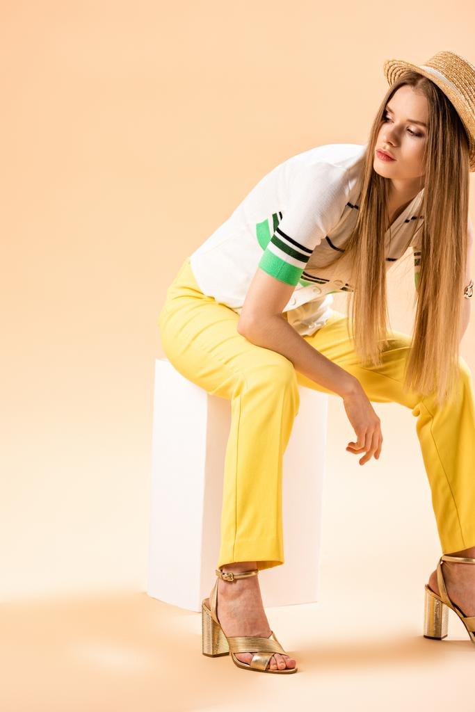 elegante ragazza in pantaloni gialli, polo e cappello di paglia seduta su cubo bianco su beige
 - Foto, immagini