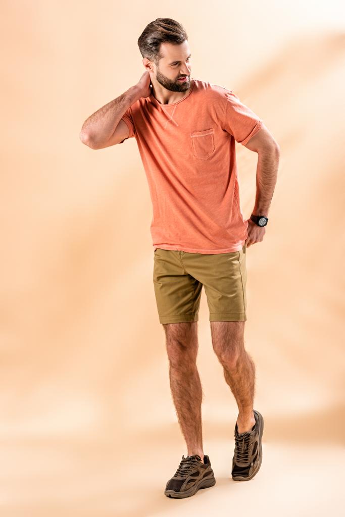 κομψός όμορφος άντρας ποζάρει με σορτς και καλοκαιρινό t-shirt σε μπεζ - Φωτογραφία, εικόνα