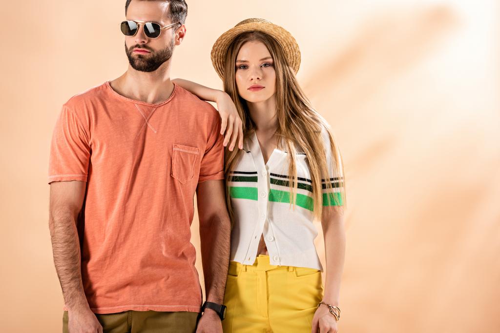 молода стильна пара позує в літньому одязі, солом'яному капелюсі і сонцезахисних окулярах на бежевому
 - Фото, зображення