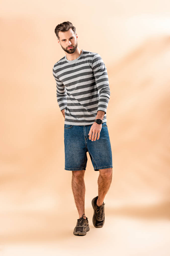 стильний бородатий молодий чоловік позує в смугастій сорочці на бежевому
 - Фото, зображення