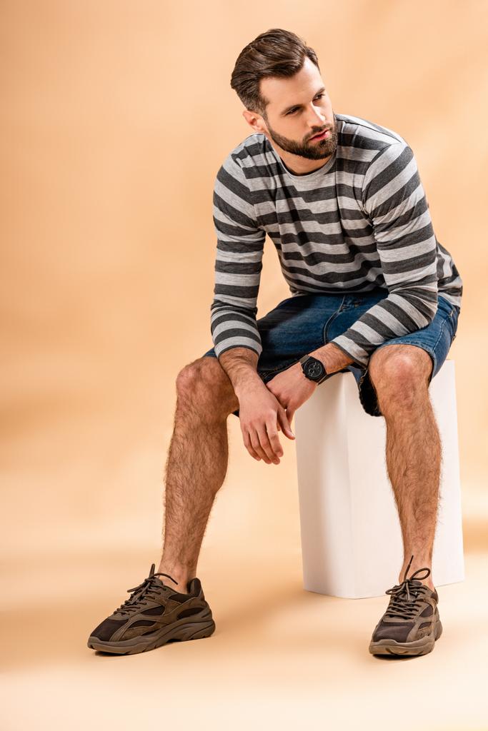 bearded man in striped sweatshirt sitting on white cube on beige - Photo, Image