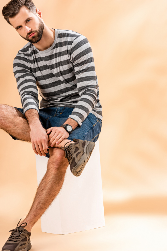 jóképű szakállas férfi csíkos pulóverben ül fehér kockán bézsen - Fotó, kép