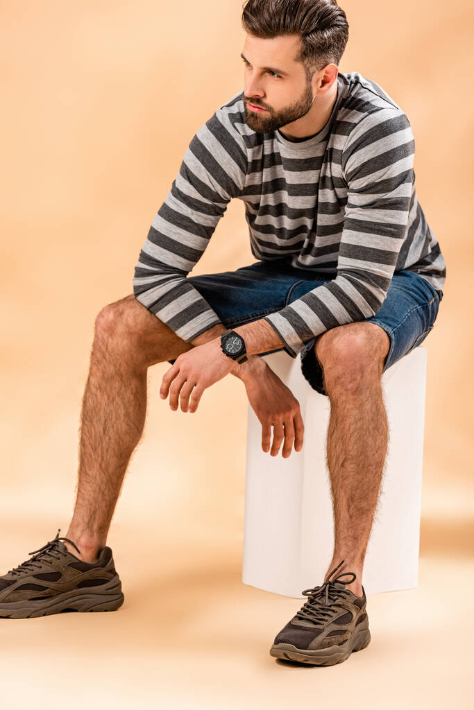 красивый стильный бородатый мужчина в полосатой толстовке сидит на белом кубе на бежевом
 - Фото, изображение