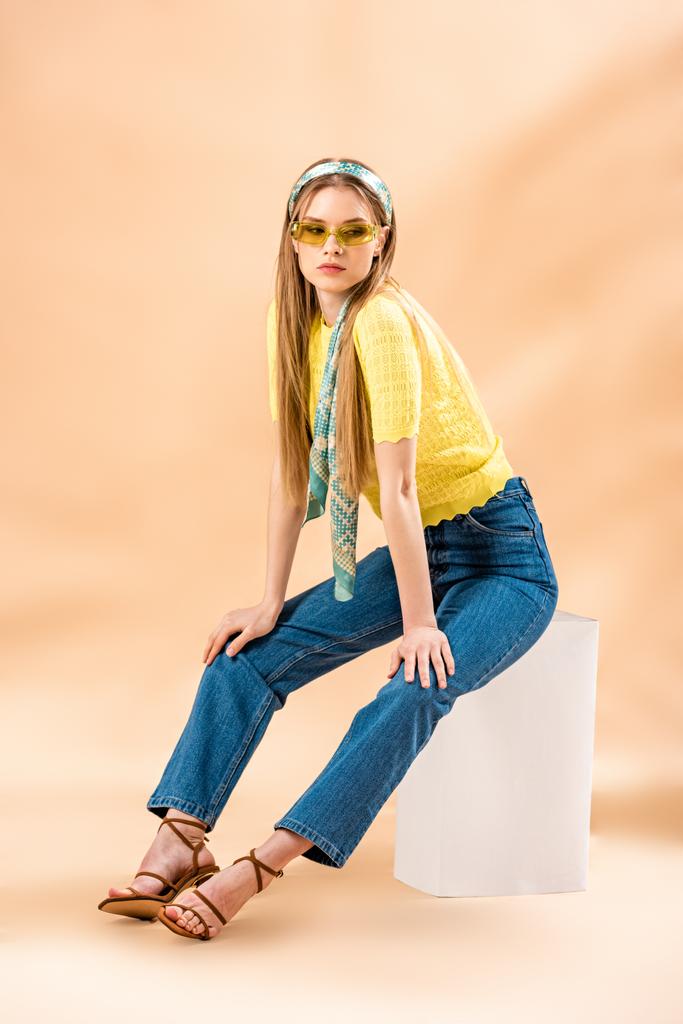 hermosa chica rubia en jeans, camiseta amarilla, gafas de sol, sandalias de tacón y bufanda de seda sentado en cubo blanco en beige
 - Foto, imagen