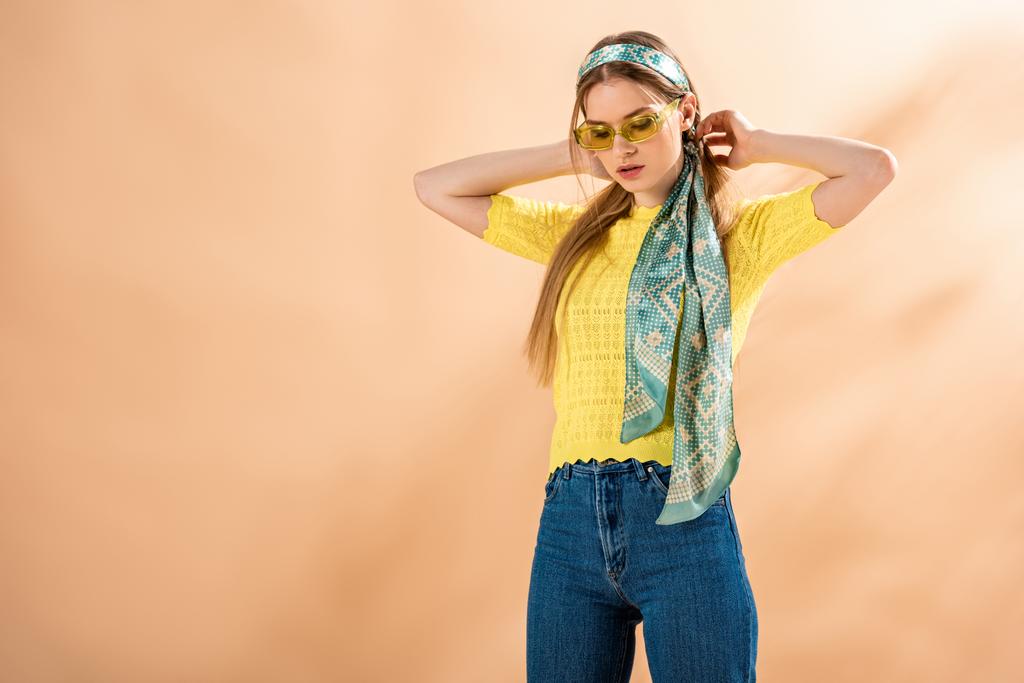 красива стильна дівчина позує в джинсах, жовтій футболці, сонцезахисних окулярах і шовковому шарфі на бежевому
 - Фото, зображення