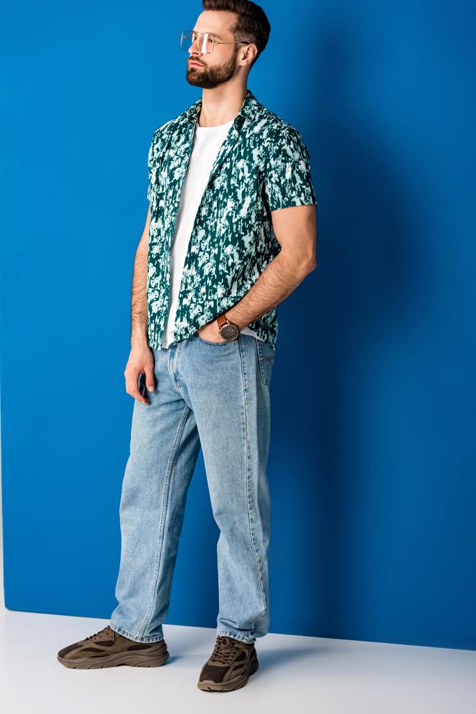 μοντέρνα γενειοφόρος άνδρας ποζάρουν σε καλοκαιρινά ρούχα και γυαλιά ηλίου σε γκρι και μπλε - Φωτογραφία, εικόνα