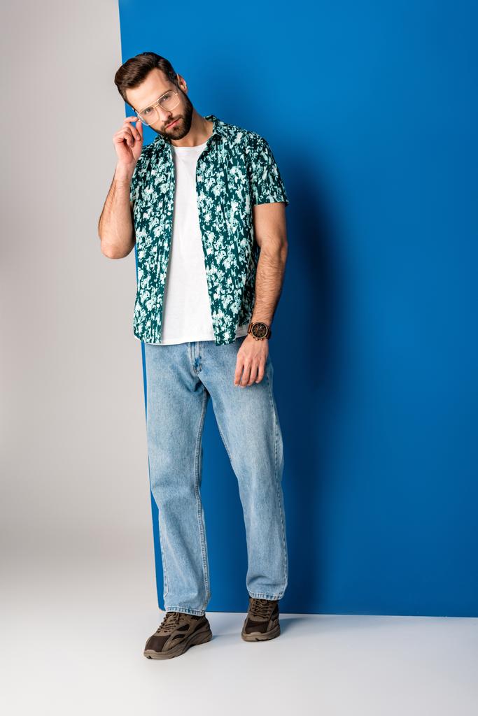 bärtiger stylischer Mann posiert in Sommerkleidung und Sonnenbrille auf grau und blau - Foto, Bild