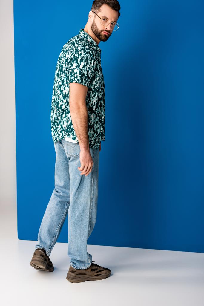 stylový vousatý muž pózující v letních šatech a slunečních brýlích na šedé a modré - Fotografie, Obrázek