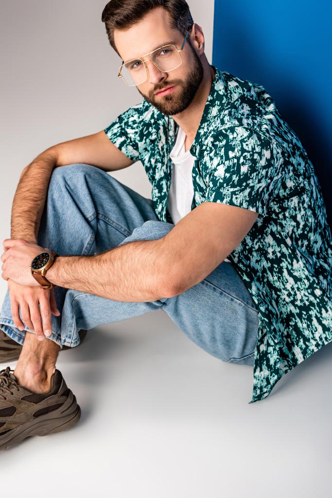 стильный молодой человек позирует в летней одежде и солнцезащитных очках на сером и синем
 - Фото, изображение