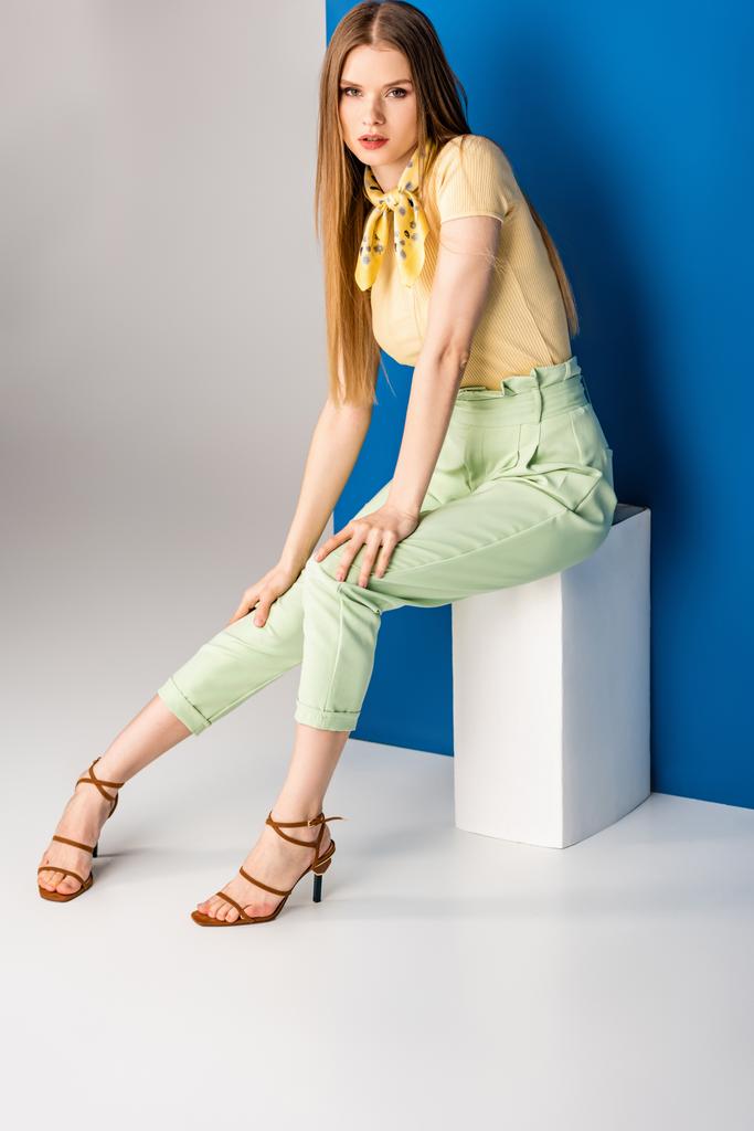 beautiful woman in green summer trousers and heeled sandals sitting on white cube on grey and blue - Photo, Image