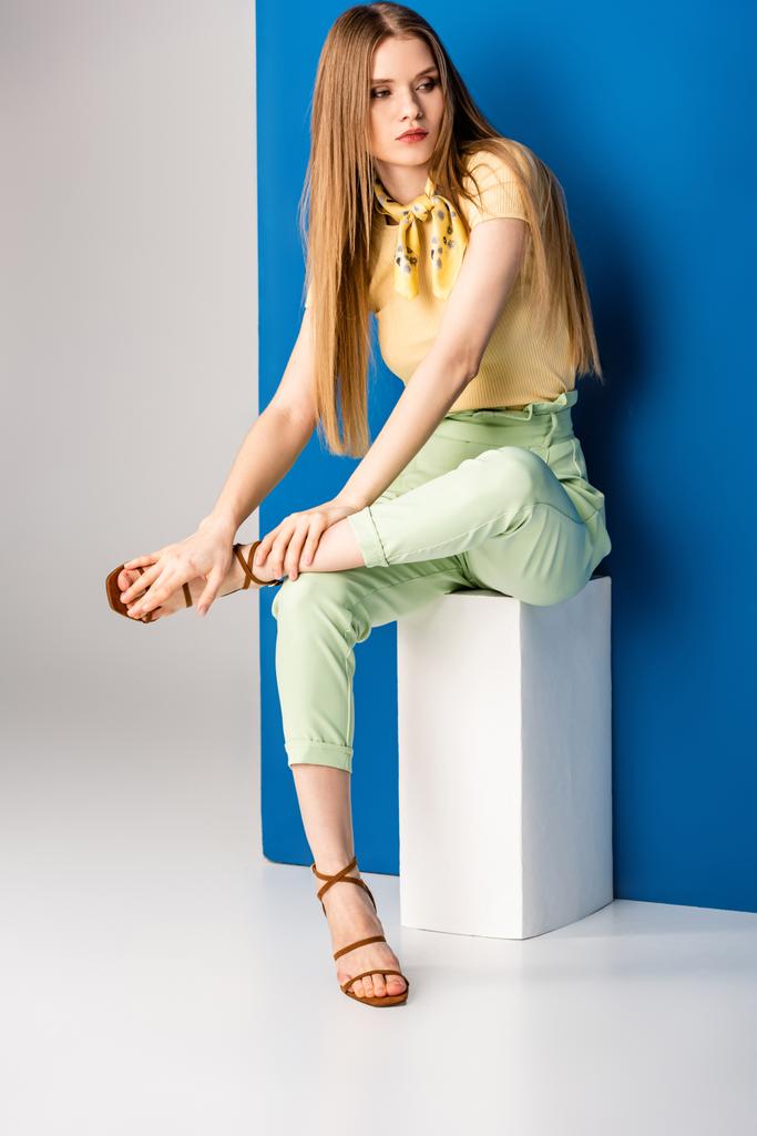 modna kobieta w zielonych letnich spodniach i sandałach na obcasach siedząca na białym sześcianie na szaro-niebieskim - Zdjęcie, obraz