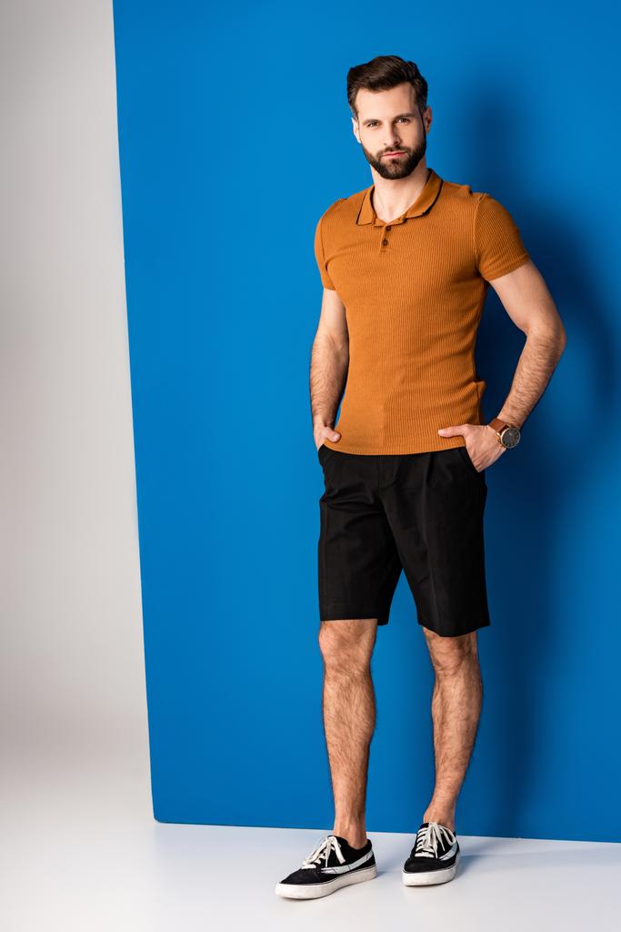 elegante barbudo joven posando en pantalones cortos y polo marrón en gris y azul
  - Foto, imagen