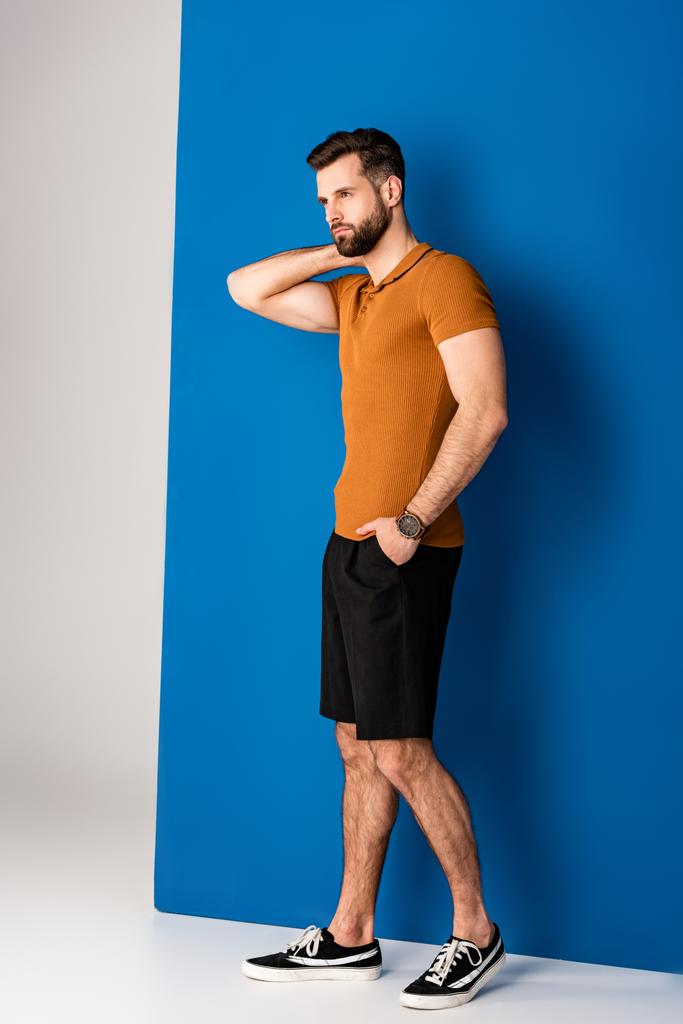 knappe modieuze man poserend in shorts en bruine polo op grijs en blauw  - Foto, afbeelding