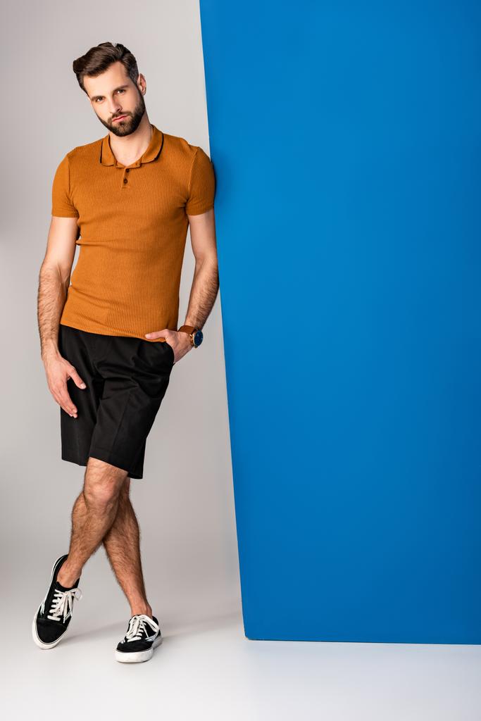 knappe stijlvolle man met baard poserend in shorts en bruine polo op grijs en blauw  - Foto, afbeelding