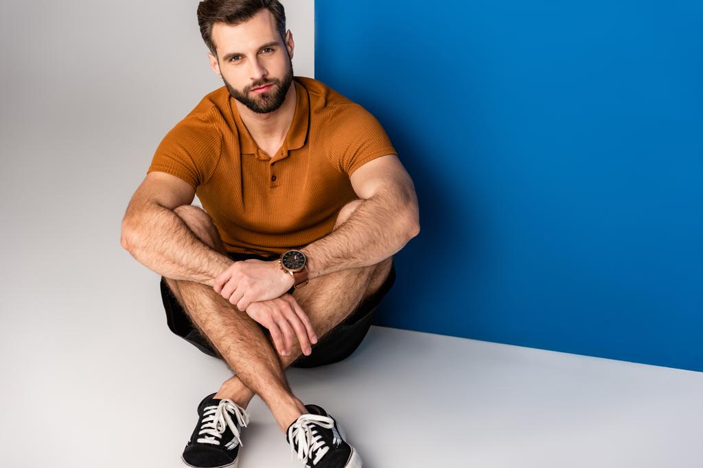 fashionable bearded man sitting in shorts and brown polo on grey and blue - Photo, Image