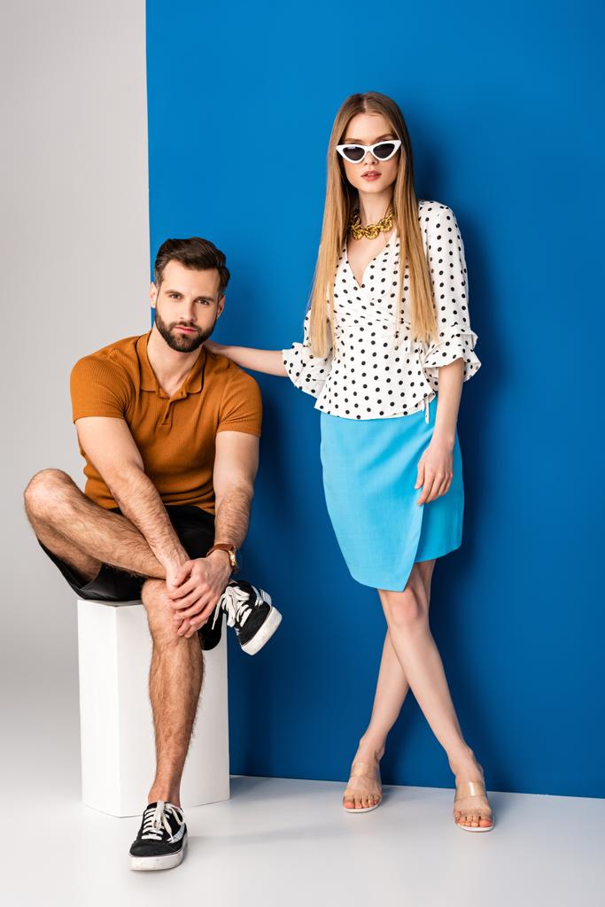 trendy jong paar in de zomer kleding en zonnebril poseren in de buurt van witte kubus op grijs en blauw - Foto, afbeelding