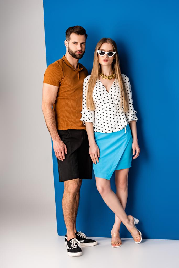 trendy paar poseren in de zomer kleding en zonnebril op grijs en blauw - Foto, afbeelding