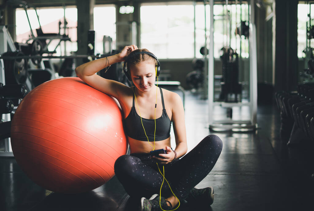 Een mooie vrouw met een sportshirt zit en ontspant door naar muziek te luisteren vanaf een koptelefoon. met rubberen yogabal of oefenbal - Foto, afbeelding
