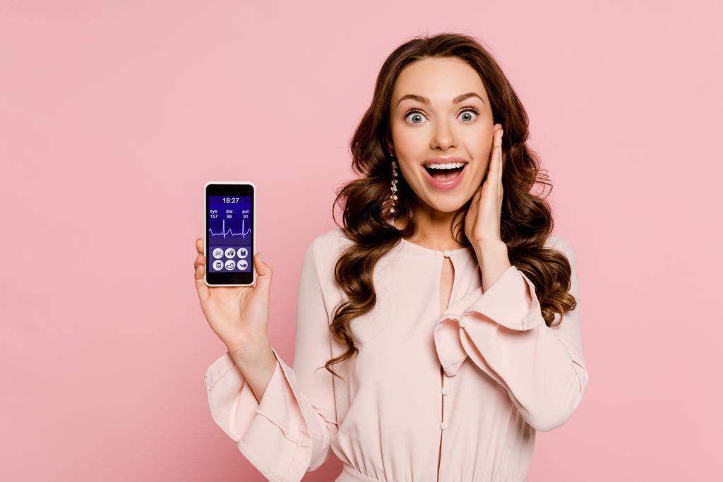 興奮した女の子が画面上に医療アプリでスマートフォンを保持し、ピンクで隔離されたカメラを見て  - 写真・画像