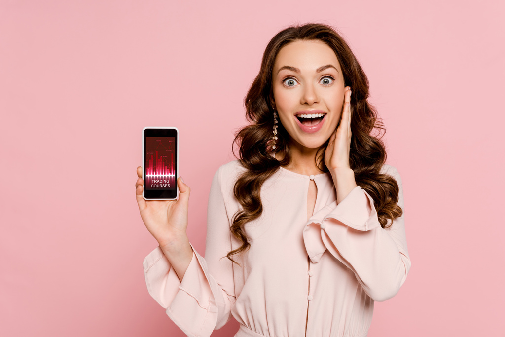 vzrušená dívka držící smartphone s obchodními kurzy na obrazovce a při pohledu na kameru izolované na růžové  - Fotografie, Obrázek