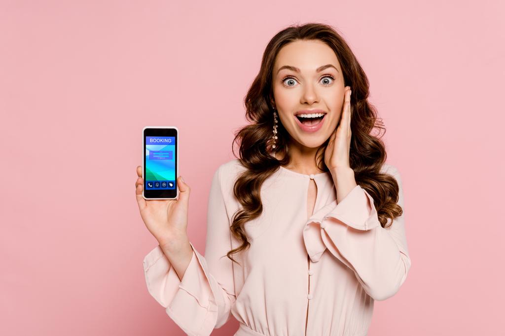 vzrušená dívka držící smartphone s rezervací na obrazovce a při pohledu na kameru izolované na růžové  - Fotografie, Obrázek