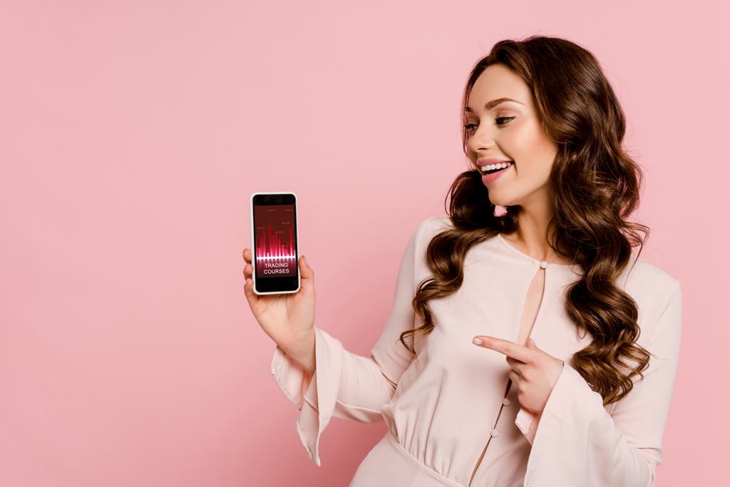 vrolijk meisje wijzen met de vinger naar smartphone met trading cursussen op het scherm geïsoleerd op roze  - Foto, afbeelding
