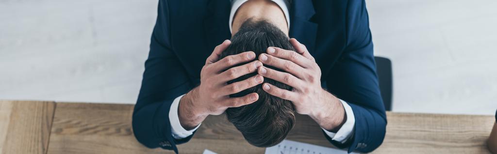plano panorámico de empresario deprimido cogido de la mano en la cabeza inclinada mientras se sienta en el escritorio en la oficina
 - Foto, Imagen