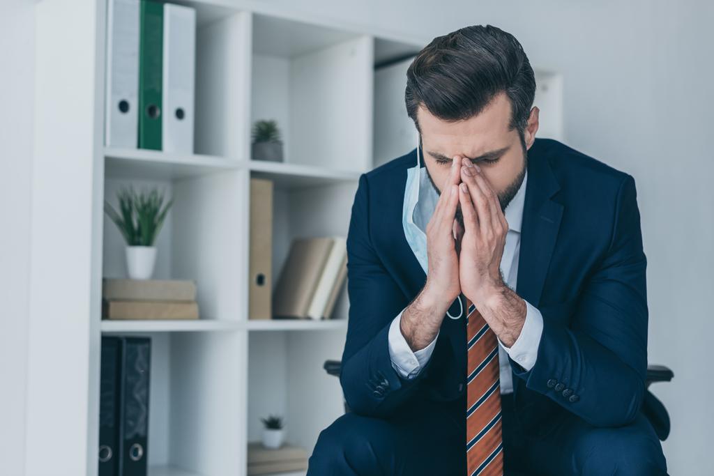 depressieve zakenman aanraken gezicht terwijl zitten in het kantoor met gebogen hoofd en gesloten ogen - Foto, afbeelding