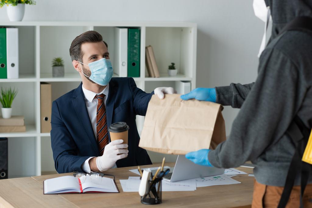 częściowy widok dostawcy żywności człowiek daje torbę papierową do biznesmena w masce medycznej w biurze - Zdjęcie, obraz
