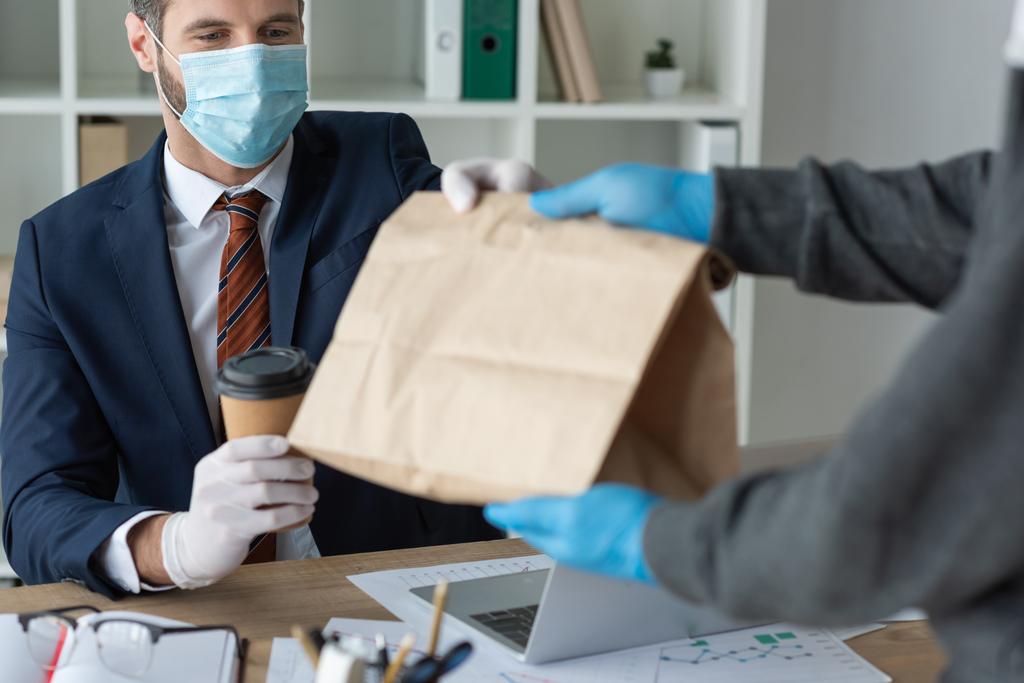 cropped view of food delivery man giving paper bag to businessman in medical mask in office - Photo, Image