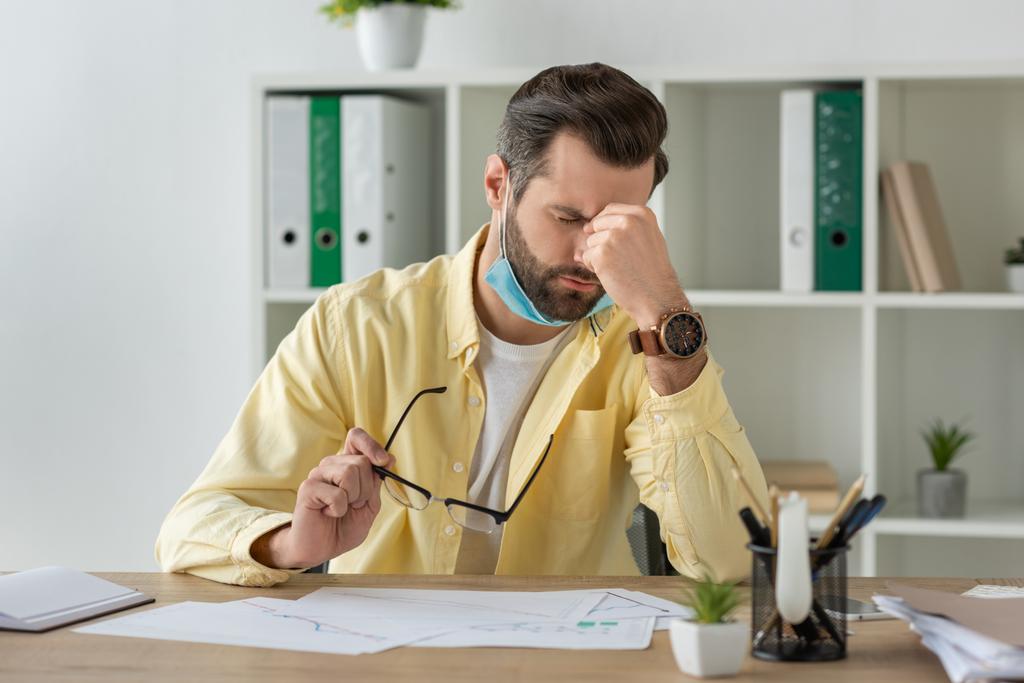 empresário exausto segurando óculos e tocando rosto enquanto sentado no local de trabalho perto de documentos
 - Foto, Imagem