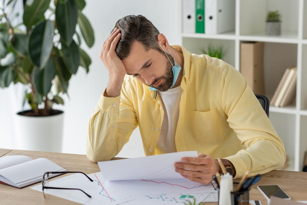 thoughtful businessman holding hand on head while sitting at workplace and looking at document - Photo, Image