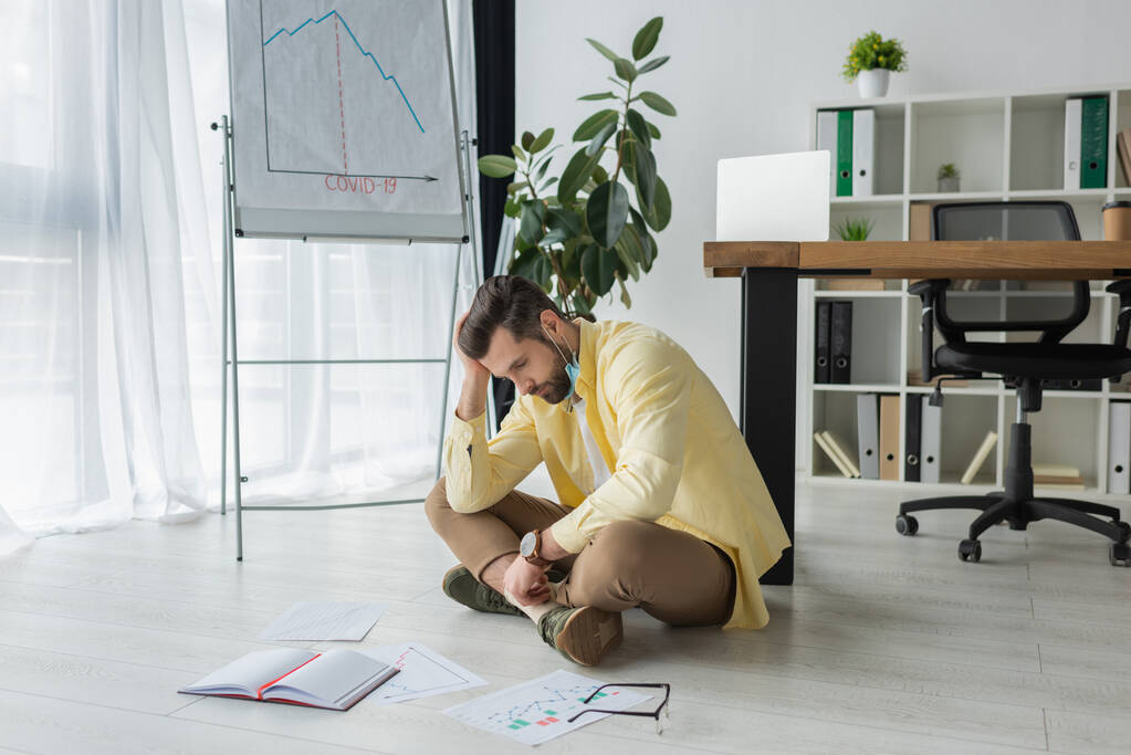 depressed businessman sitting on floor near papers, notebook and flipchart with covid-19 inscription and graphs showing decrease - Photo, Image