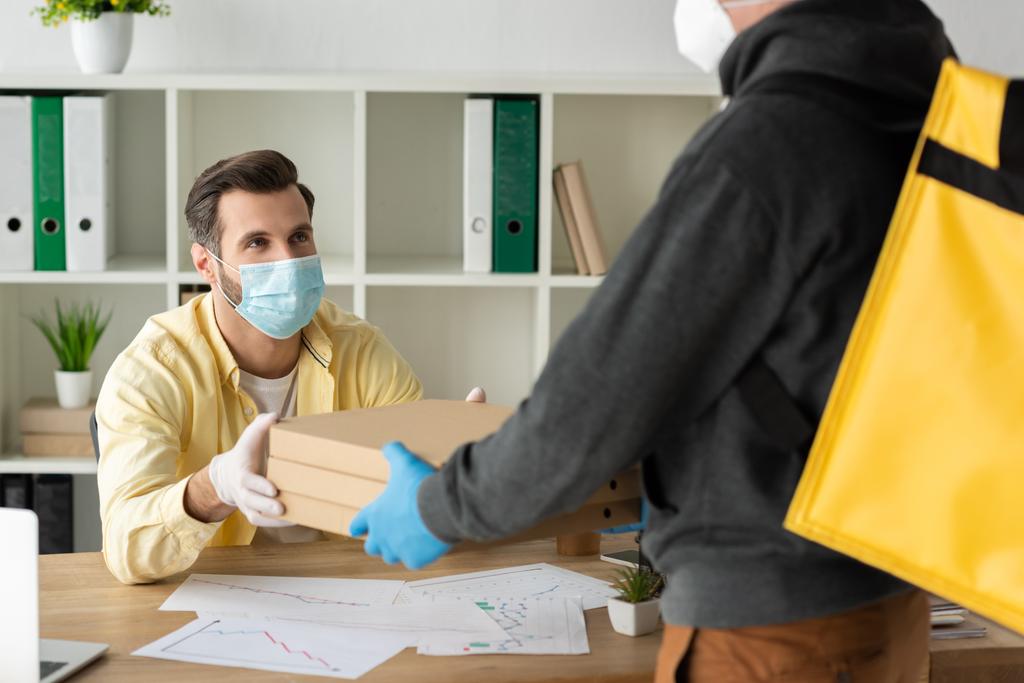 vista parcial del repartidor de comida hombre dando cajas de pizza a hombre de negocios en máscara médica y guantes de látex
 - Foto, Imagen