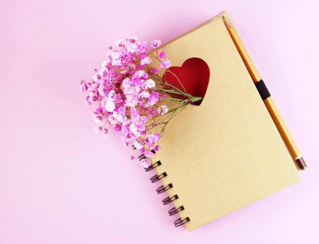 hnědý zápisník s květinami na růžovém pozadí. Zápisník s červeným srdcem a tužkou na pastelovém pozadí. fialová jarní kytice s notebookem - Fotografie, Obrázek