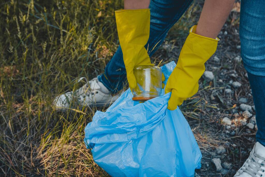 Μια νεαρή γυναίκα μαζεύει τα σκουπίδια και βάζει σε μια πλαστική σακούλα σκουπιδιών - έννοια της περιβαλλοντικής ρύπανσης - Φωτογραφία, εικόνα