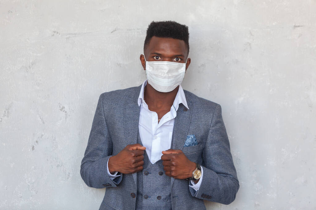 COVID-19. Un joven africano de confianza lleva una mascarilla médica protectora para prevenir la infección con coronavirus y pandemia de pie en un traje gris contra una pared de hormigón.
 - Foto, Imagen