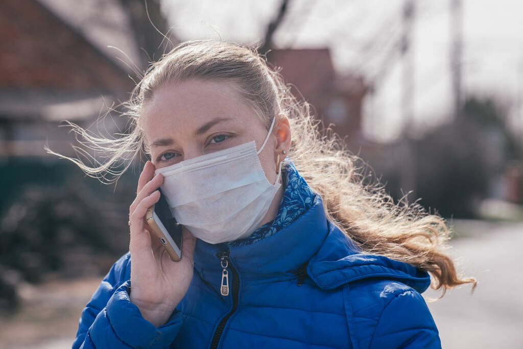 Λευκή κοπέλα σε αναρρωτική άδεια με ιατρική προστατευτική μάσκα, τηλεφωνεί στο τηλέφωνο, καλεί ταξί. Εποχιακή γρίπη και επιδημία κρυολογήματος. Προστασία από αναπνευστικές νόσους κατά τη διάρκεια πανδημιών και επιδημιών - Φωτογραφία, εικόνα