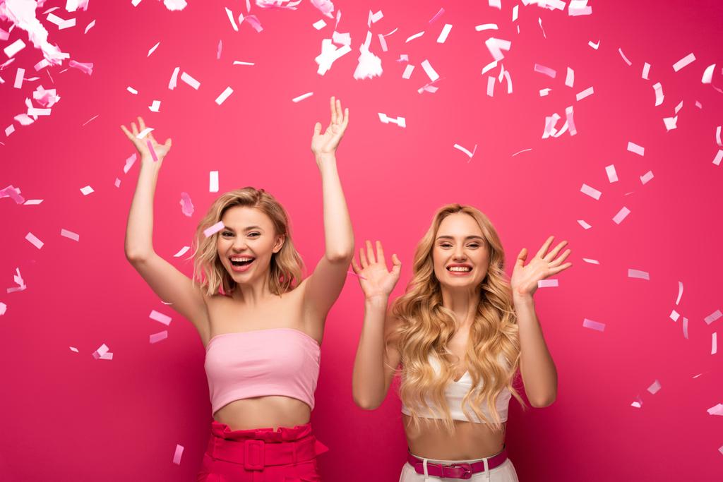 Cheerful blonde friends smiling at camera under falling confetti on pink background - Photo, Image