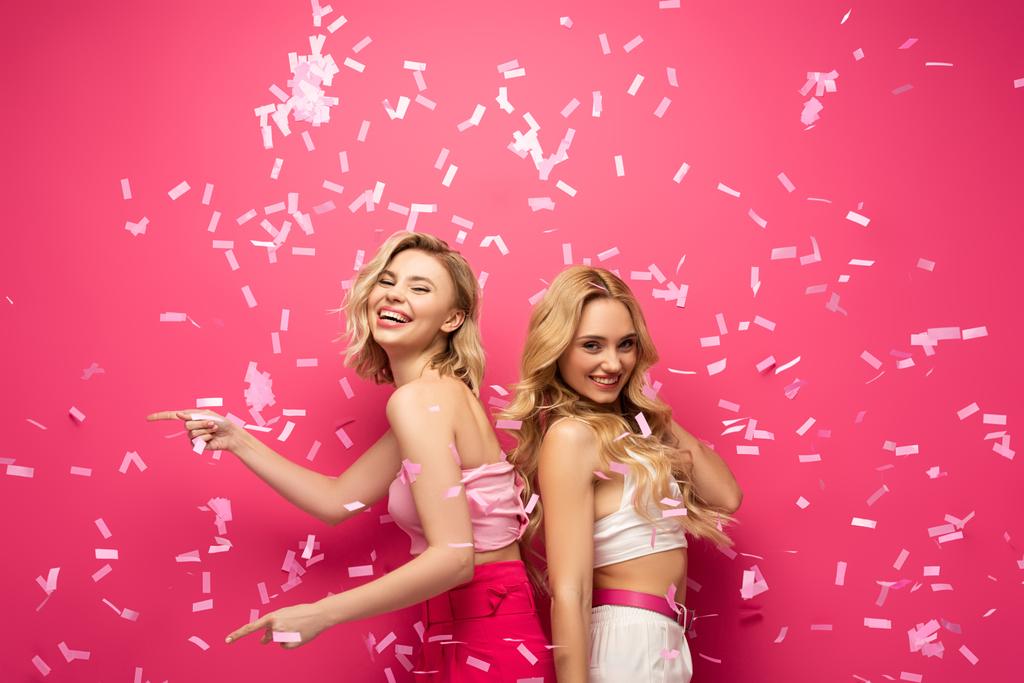 Zijaanzicht van glimlachende blonde vrienden kijken naar de camera en wijzen met vingers onder vallende confetti op roze achtergrond - Foto, afbeelding