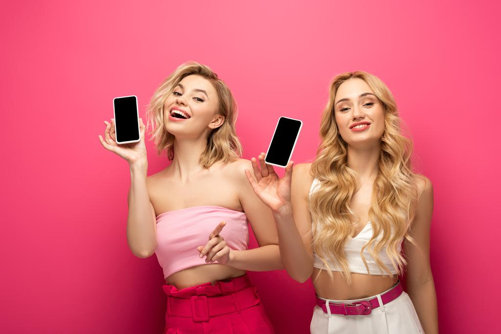 Усміхнені блондинки тримають смартфони з порожніми екранами на рожевому фоні
 - Фото, зображення