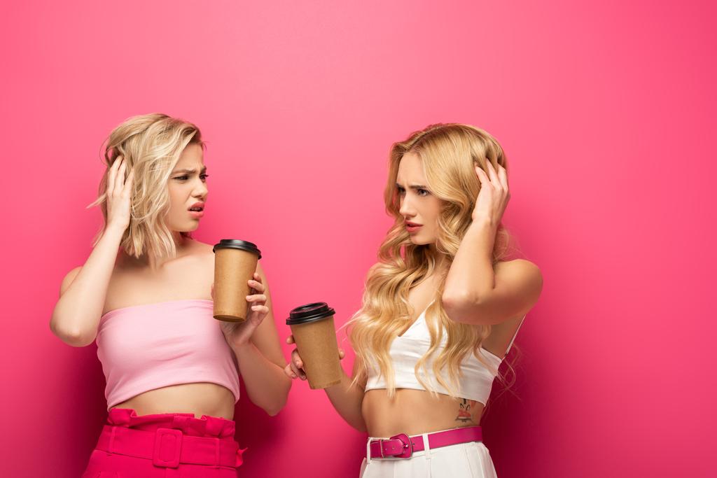 Pensive blonde sisters holding coffee to go on pink background - Photo, Image