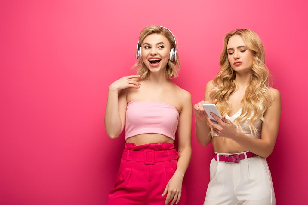 Veselé blondýny dívka ve sluchátkách v blízkosti přítele pomocí smartphone na růžovém pozadí - Fotografie, Obrázek