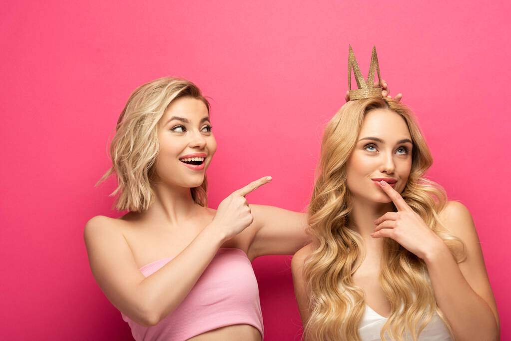 Smiling blonde woman pointing on pensive friend in crown on pink background - Photo, Image