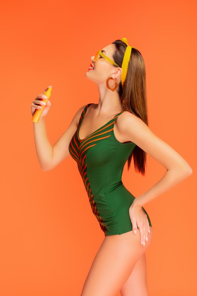 Вид збоку жінки з рукою на стегно посміхається і наносить сонцезахисний крем ізольований на апельсин
 - Фото, зображення